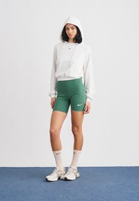 Γκρι φούτερ με κουκούλα και λευκά κορδόνια, πράσινα σορτς, μπεζ αθλητικά παπούτσια και εκρού κάλτσες με μαύρες λεπτομέρειες, με λογότυπο της Nike.