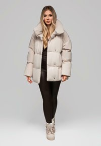Béžová péřová bunda se zvýšeným límcem, oversize střihem, patentkami a prošívaným designem. Černé legíny a béžové vysoké tenisky.