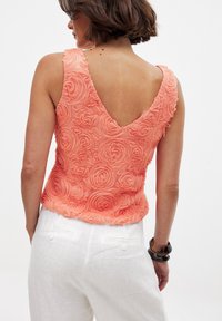 Top senza maniche color corallo con schiena a V, caratterizzato da una texture in rilievo a motivo di rose. Abbinato a pantaloni di lino bianchi e a un bracciale scuro.