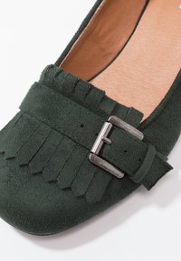 Chaussure plate en daim vert avec un détail à franges, une boucle métallique en accent et une doublure intérieure lisse couleur tan. Texture douce sur toute la surface.