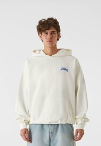 Krēmkrāsas kapuce, izgatavota no mīksta auduma, ar priekšējo kabatu un zilu grafiku kreisajā krūtīs. Brīvs griezums ar ribotām piedurknēm.