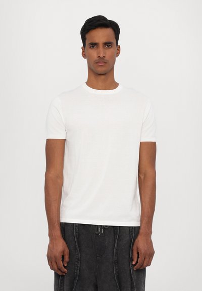 T-shirt blanc à manches courtes avec un col rond. Tissu doux et léger, légèrement ajusté, porté avec un pantalon sombre et texturé.