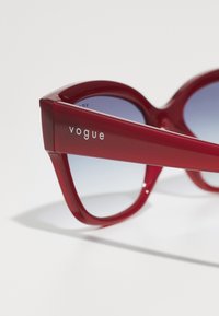 Ochelari de soare din plastic roșu cu un design unghiular îndrăzneț, lentile din gradient albastru și logo-ul "vogue" în argintiu pe braț.