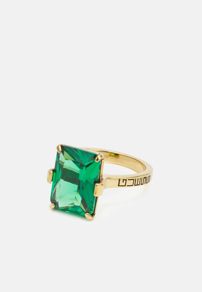 Bague en or avec une grande pierre précieuse verte rectangulaire et une gravure géométrique noire sur l’anneau, sur un fond blanc uni.