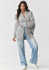 Szary sweter z dzianiny o fakturze warkocza, z otwartym przodem i oversize'owymi rękawami, zestawiony z jasnoniebieskimi spodniami typu wide-leg i białymi sneakersami.