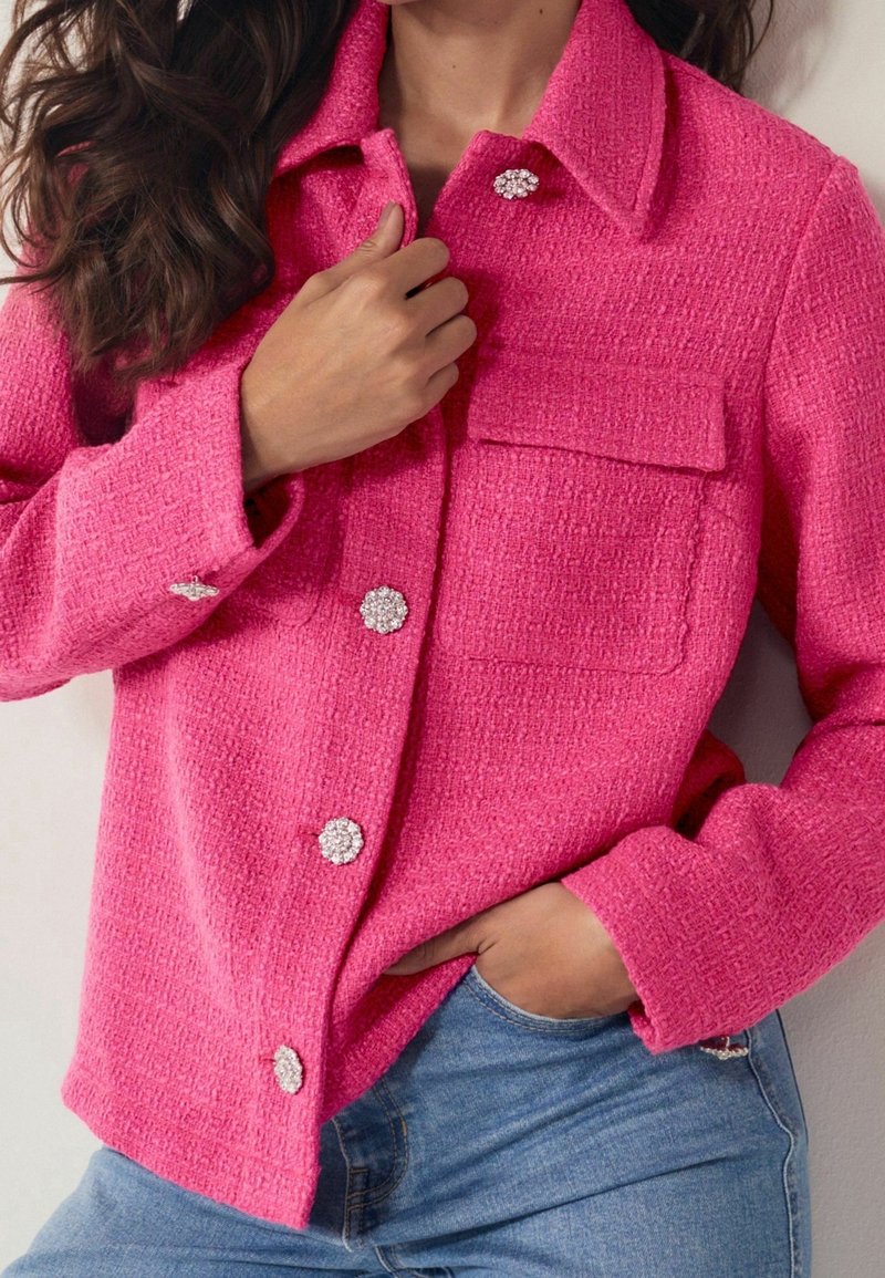 Femme portant une veste texturée rose vif avec de gros boutons en strass, une main ajuste le col, l'autre main dans la poche d'un jean bleu.