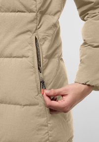 Beige vadderad jacka med quiltad textur, utrustad med en sidodragkedja och svart och orange dragsko. En hand syns justera dragkedjan.