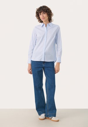 Femme aux cheveux bouclés portant une chemise bleu clair boutonnée, un jean bleu large et des baskets blanches, debout devant un fond uni.