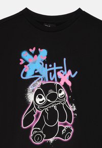 Camiseta negra con un gráfico de un personaje de dibujos animados con una expresión juguetona, rodeado de corazones rosas y acentos de pintura en spray azul.