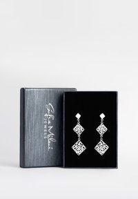 Silverhängande örhängen med intrikata utskärningsmönster, som har diamantformer och två hängande sektioner, visade i en svart smyckeslåda.