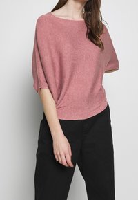 Kortärmad, löst sittande topp i strukturerad rosa stickad tyg; har en vid halsringning och en lätt fåll med subtila horisontella linjer.