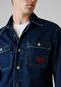 Camisa de denim en azul oscuro con dos bolsillos en el pecho, botones a presión plateados y texto bordado en rojo "RED BULL RACING". Detalles de costura visibles.