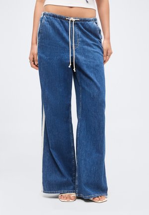 Bredbenede blå denim bukser med hvid snor i taljen, siderstriber, båret med hvide sandaler, viser mave og hænder i siderne.