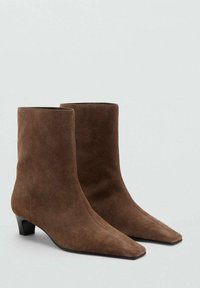 Botas de ante marrón hasta el tobillo con tacones bajos, punteras afiladas y un diseño suave y sin costuras sobre un fondo neutro.