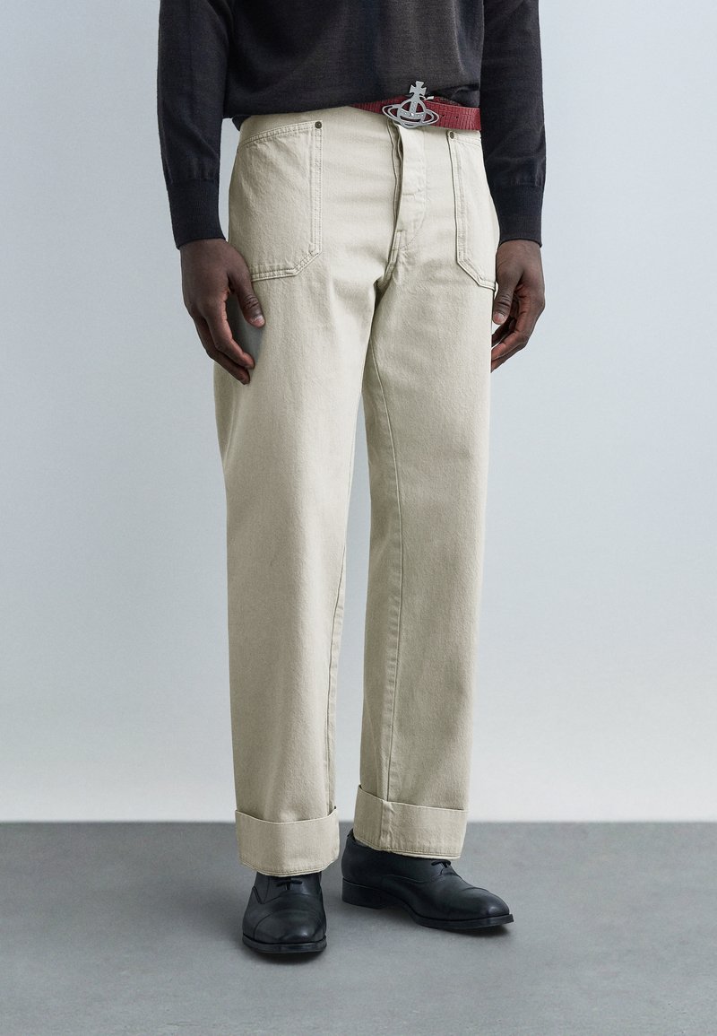 Άνδρας που φοράει μπεζ παντελόνι με φαρδιά ίσια πόδια και μανσέτες, κόκκινη ζώνη με ασημένια αγκράφα, μαύρα παπούτσια και σκούρο πουκάμισο μακρυμάνικο, στέκεται σε γκρι δάπεδο.