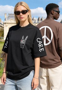 Jeune femme avec lunettes de soleil noires et t-shirt montrant le symbole de la paix avec la main et des mots, et homme en t-shirt marron avec un grand signe de paix dans le dos en extérieur.