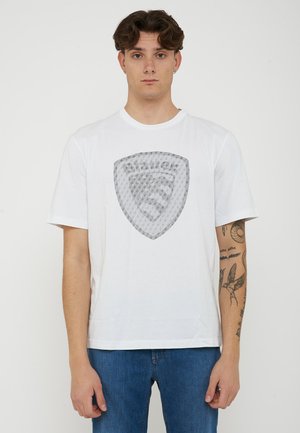 Giovane uomo con capelli castani corti che indossa una maglietta bianca con un motivo a scudo e jeans blu, in piedi davanti a uno sfondo uniforme.