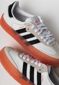 Białe i szare skórzane sneakersy z czarnymi paskami, pomarańczową przezroczystą podeszwą i teksturowanymi akcentami, z brandingiem "Samba".