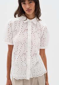 Blusa bianca ricamata con maniche corte, chiusura con bottoni sul davanti, intricate applicazioni a intaglio e orlo scallop.