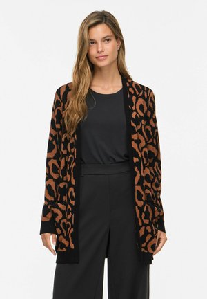 Femme aux longs cheveux portant un haut noir et un pantalon, ainsi qu'un cardigan ample marron et noir à motif léopard.