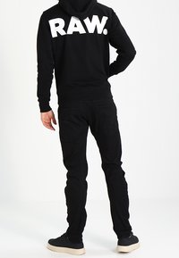 Black hoodie with large white "RAW." text on back, paired with black pants and black sneakers with white soles. Casual outfit viewed from behind.