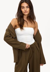 Vrouw met lang bruin haar, gekleed in een witte strapless top en een losse bruine broek met een ongepaste schouderjacket, met haar handen op de taille.