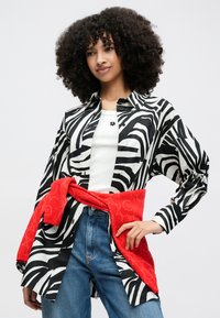 Μπλούζα με ασπρόμαυρες ρίγες από ελαφρύ ύφασμα, συνδυασμένη με κόκκινο πουλόβερ δεμένο γύρω από τη μέση πάνω από τζιν παντελόνι.