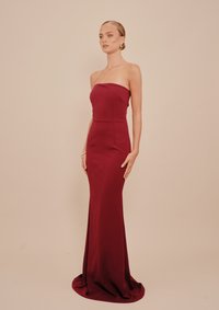 Γυναίκα που στέκεται φορώντας ένα strapless, μακρύ μπορντό φόρεμα με εφαρμοστή κορυφή και ελαφριά ουρά, σε απλό μπεζ φόντο.