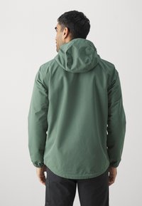 Veste légère verte avec capuche, poignets élastiques et texture lisse. Le design est simple et fonctionnel sans matériel visible.