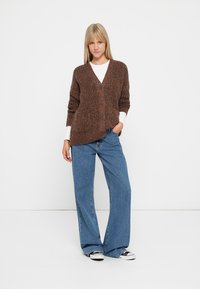 Brązowy sweter o fakturowanym splocie z dekoltem w serek, zapinany na guziki, z dwiema przednimi kieszeniami, zestawiony z jasnoniebieskimi spodniami typu wide-leg i czarnymi trampkami.