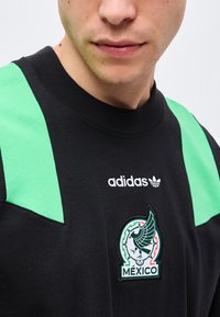 Gros plan d'une personne portant un sweat-shirt Adidas noir avec des panneaux verts sur les épaules et un emblème du Mexique représentant une figure ailée et un ballon de football.