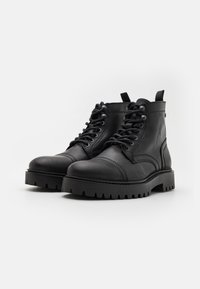 Botas de cuero negras con punta redondeada, diseño de cordones y costuras en relieve. Cuentan con una suela gruesa y estriada y una lengüeta en el talón.