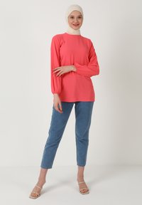 Maglietta rosa a maniche lunghe con polsini arricciati, abbinata a jeans blu con risvolto e sandali chiari. Il modello indossa un hijab color crema. Outfit semplice e informale.