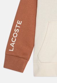 Felpa con cappuccio caratterizzata da un corpo crema e una manica color ruggine, con la scritta "LACOSTE" stampata in bianco. Realizzata in un tessuto morbido, include una tasca frontale.