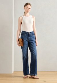 Débardeur crème, jean en denim bleu taille haute, pochette marron et sandales marron à lanières. Sol en bois naturel avec un arrière-plan neutre.