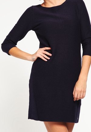 Woman wearing a fitted black dress with three-quarter sleeves, one hand on hip and a small anchor tattoo on wrist visible.