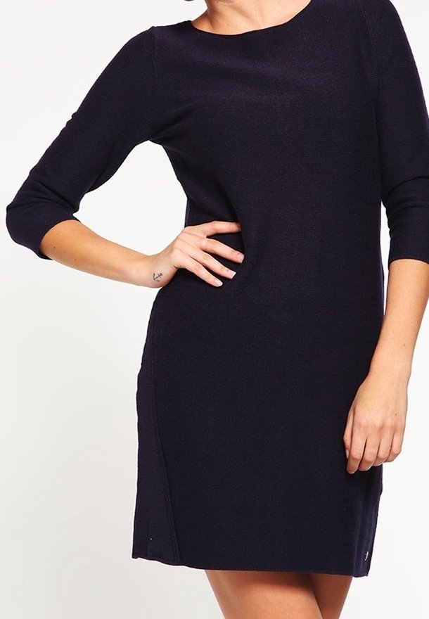 Femme portant une robe noire moulante avec des manches trois-quarts, une main sur la hanche et un petit tatouage d’ancre visible sur le poignet.