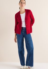 Roter, gerippter Cardigan mit V-Ausschnitt und fünf Knöpfen, getragen über einem weißen Oberteil und kombiniert mit weit geschnittenen blauen Jeans und weißen Sneakern.