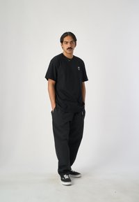 Schwarzes Baumwoll-V-Ausschnitt-T-Shirt mit kleinem Logo auf der Brust, kombiniert mit locker sitzenden schwarzen Hosen und schwarzen Sneakers mit weißen Sohlen.