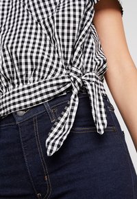 Blusa a cuadros en blanco y negro con cintura fruncida y detalle atado, combinada con unos jeans de mezclilla oscuros con cierre de botón y costuras doradas.