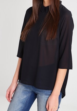 Vrouw draagt een losse zwarte blouse met driekwart mouwen en een blauwe spijkerbroek, staand tegen een effen witte achtergrond.