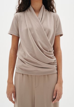 Kobieta ubrana w beżowy top z krótkim rękawem z marszczonym przodem oraz dopasowane luźne spodnie, stojąca na tle białego, jednolitego tła.