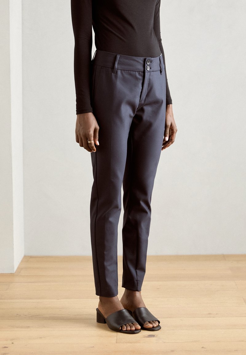 Navy trousers with a slim fit, two-button closure, and side pockets, paired with black slide sandals and a black long-sleeve top.