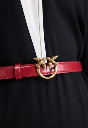 Ceinture en cuir rouge avec boucle dorée ornée de deux oiseaux, portée sur un tissu noir et blanc.