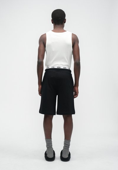 Schwarze Shorts mit einem kontrastierenden elastischen Bund, der ein Logo trägt. Kombiniert mit einem weißen Tanktop, grauen Socken und schwarzen Schuhen.