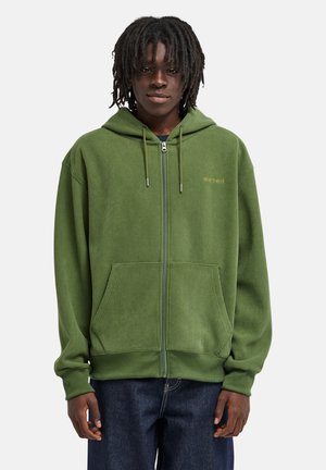 Giovane con dreadlocks che indossa una felpa verde con zip e jeans scuri, in piedi davanti a uno sfondo chiaro e semplice.