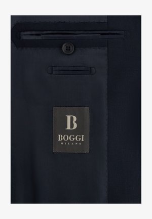 Tummansinisen puvun sisäpuoli, jossa näkyvä nappi, ommellut taskut ja "Boggi Milano" -merkkilappu.