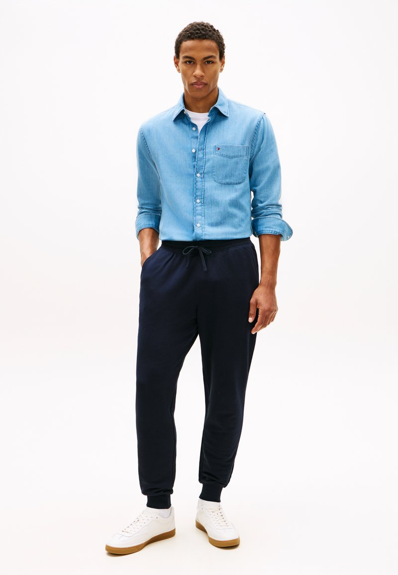 Uomo con camicia azzurra chiara abbottonata con maniche arrotolate, pantaloni da jogging scuri e sneakers bianche, in piedi con le mani in tasca.