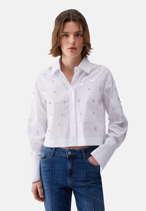 Giovane donna che indossa una camicia bianca con bottoni e decorazioni argentate e jeans blu a vita alta, in piedi con una mano in tasca.