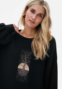 Czarna bluza z długim rękawem, z centralnym haftowanym motywem drzewa i korzeni na beżowym okręgu. Miękka tekstura.
